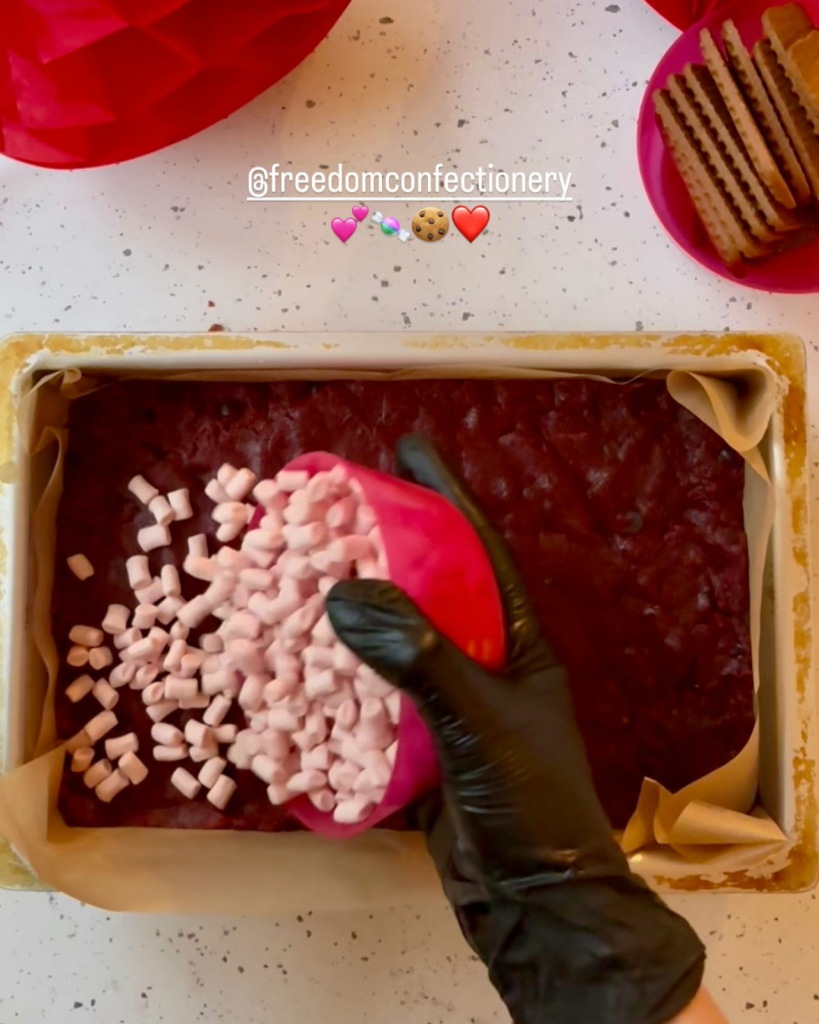 A hand in a black glove pours pink mini marshmallows onto a dark red velvet batter in a parchment-lined baking pan.