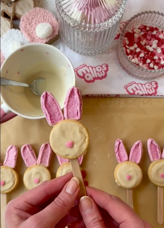A person holding a finished vegan Easter bunny lolly made with a biscuit, pink ears, and a heart nose. More lollies are on the table.